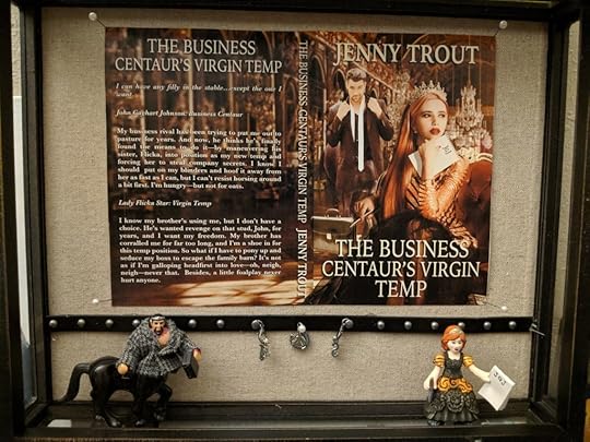 A shadow box containing a cover flat (described later) for The Business Centaur's Virgin Temp by Jenny Trout, the two action figures standing beneath it, and various charms hanging from push pins on a strip of studded leather across the lower quarter of the box.