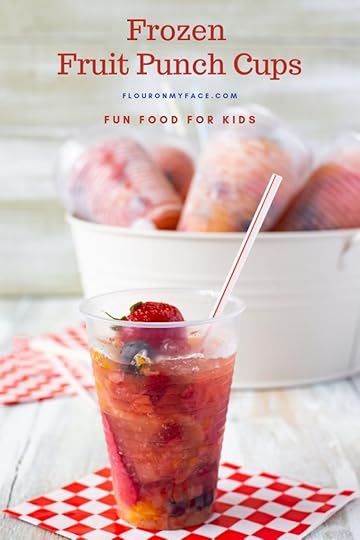 A clear plastic cup filled with fruit and punch then frozen. With a metal bucket in the background filled with ice and more Frozen Fruit Punch Cups chilling for a summer frozen treat. for the Fourth of July