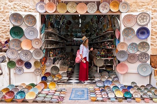 Person in long red skirt, hat and with pink backpack is standing in a small shop and looking at Moroccan colorful plates