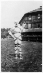 A mummy-like figure standing in front of a barn, 1919