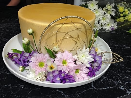 Front shot of cake, a single-tier 23cm round cake covered in fondant (with sharp corners!), sprayed gold, with a glittery gold dream catcher centered on the front side of the cake (it extends above the edge of the cake by about a centimetre); three sprays of purpley-blue wisteria are arranged around the bottom of the cake at the front on the white platter; they are filled out with three baby pink daisies, three white daisies, some white daisy buds, a few wisteria leaves, and a gold glittery feather or leaf made to match the dreamcatcher.