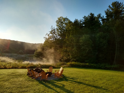 A fire pit surrounded by wooden chairs, with a small pond in the background