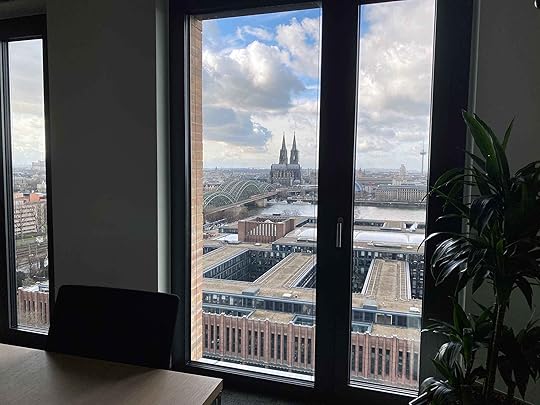 Durch ein Fenster sieht man den Rhein mit Hohernzollernbrücke und Dom. Im Vordergrund erahnt man einen Tisch mit Stuhl sowie eine Zimmerpflanze.