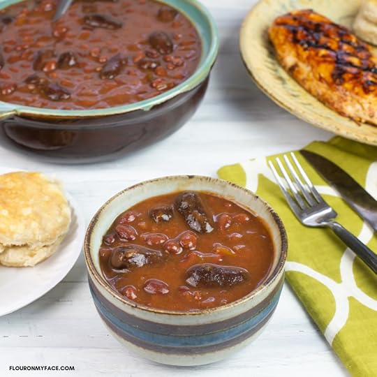 A serving bowl of Crock Pot Cowboy Beans in a small serving bowl on a table with a plate of grilled chicken, corn and a biscuit.