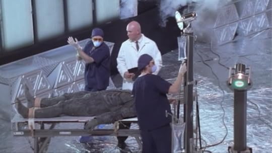 There are two people in surgical scrubs, cap, gloves, and masks working on a patient alongside a doctor in regular ass clothes with a white lab coat on, no gloves, cap, mask, anything. For some reason, the demon on the table has an IV.