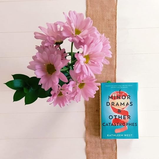 The book Minor Dramas and Other Catastrophes by Kathleen West and a bouquet of pink flowers in a vase on a white table. 