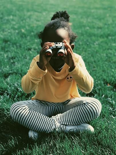 Child in yellow shirt and black and white striped pants sitting cross legged in green grass holding binoculars to eyes