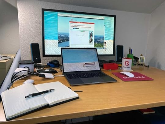 Ein Schreibtisch, darauf ein großer Monitor, davor ein Laptop. Im Bildvordergrund ein Notizbuch mit Stift. Im Hintergrund einige Kabel und eine Bonn.digital-Tasse.