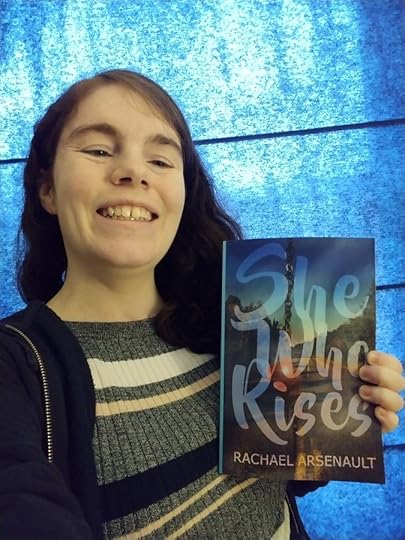 A smiling woman with dark, curly hair holds up a paperback book. The book cover features a road stretching forward in as the background, with a slightly glowing chain necklace bearing a dark red stone in the foreground. Overlaying the whole image in large, fancy letters is the title: She Who Rises. At the bottom, in smaller, simpler print, is the author name: Rachael Arsenault