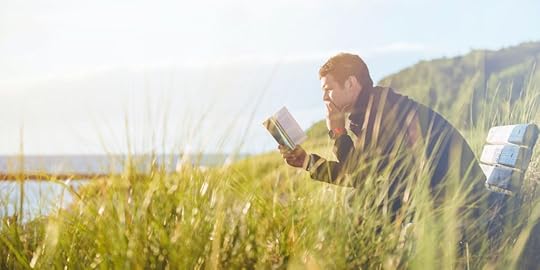 man reading a book thoughtfully, on a bench in grass by the shore.<br />