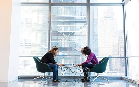 Two women work at a table with their laptops in front of large glass windows.