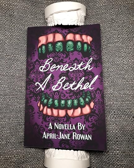 Beneath a Bethel, photo of the book with dental casts seemingly biting down on the book. The cover shows the title, white curly font, on a gothic patterned purple wallpaper-like background. Above and below the title there is a set of dentures, the teeth dark green and painted intrinsically with vines and flowers in a lighter green.