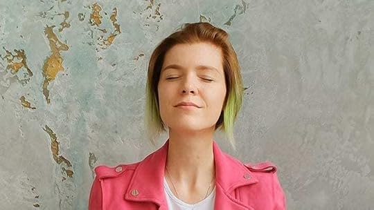 Woman with green-tipped hair and bright pink jacket meditating against a gray wall