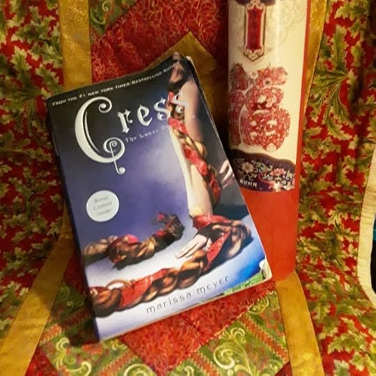 Paperback of Cress by Marissa Meyer, which features a girl's bare arm twisted up in her very long braid. The book sits on a Christmas-themed background, next to a cylinder with the Chinese character fu (blessing)