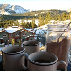 three mugs with a jar of cocoa mix in front of snow covered chalet