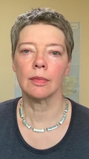 Head-and-shoulder shot of a short-haired white woman, wearing a short silver necklace and long-sleeved t-shirt, looking serious n front of a y ellow wall with a large map of early-medieval Britain on the wall behind her