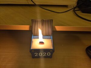 Lit candle in a small metal model of a dumpster, with the text 