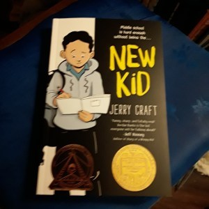 New Kid--by Jerry Craft<br /><br />The book sits on a chair<br /><br />On the cover, a kid in a gray hoodie and black slacks scribbles in a notebook