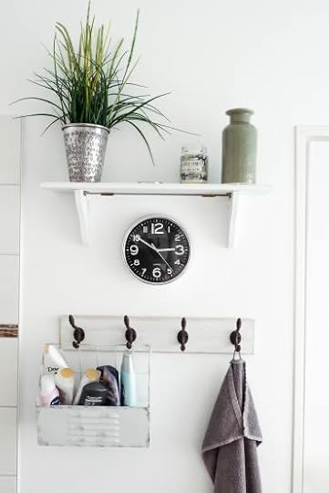 A white wall in a bathroom. On this wall is a shelf, a clock, and four hooks with bathroom essentials hung on them.