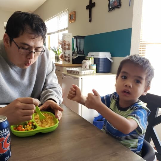 Father with dark hair blowing on mac n cheese in child's place, while toddler in green-striped shirt signs for