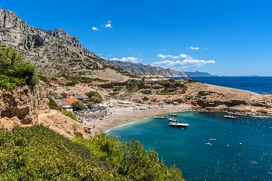 Boats floating on the blue ocean by an island with a tall mountain under a blue sky. People are mingling on the beach.