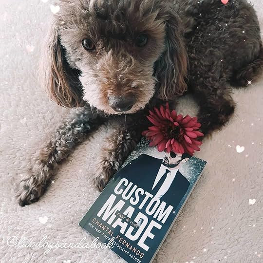A cute brown miniature poodle with a copy of the book entitled Custom Made by Chantal Fernando and a pink silk flower