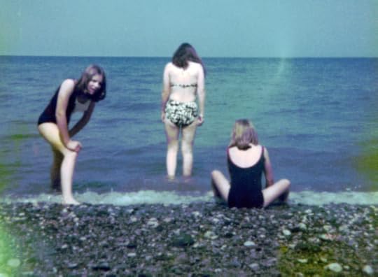 Colour photo of three girls on a beach, two in black swimsuits looking at each other, a third, in bikini staring at the horizon