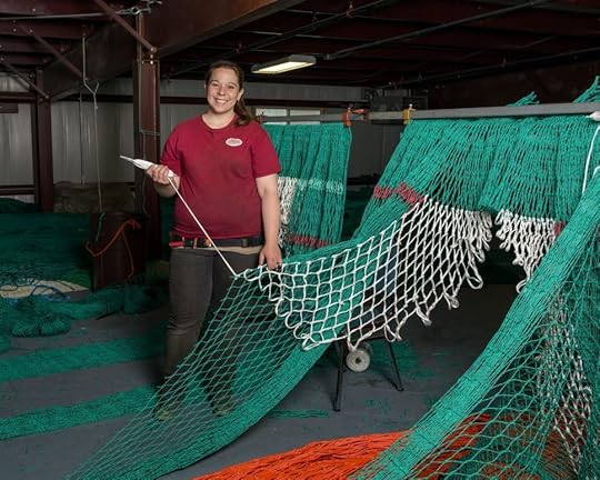 Woman stands next to a large fishing net that she is repairing. 