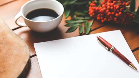 Sobre uma mesa de madeira está, no topo, uma xícara branca com café, algumas frutinhas vermelhas ao lado direito com folhas verdes ao lado. Na parte de baixo, está uma folha em branco uma caneta vermelha sobre ela.