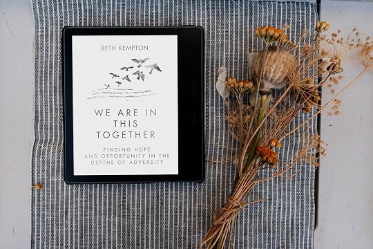 flat lay image of a tablet computer with the cover of We Are In This Together e-book displayed, next to dried bunch of flowers, on a striped cloth