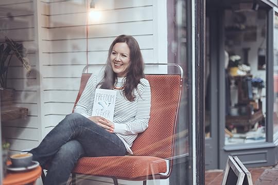 author sitting in chair laughing, holding Freedom Seeker book in hand, coffee on table in front of her, door behind her