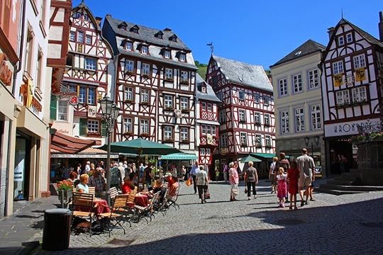 A Bavarian village square with crowd of locals. Chris is waiting while Marlowe buys them lunch. Sausages, beer, and perhaps a little change.