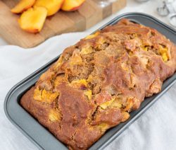 Loaf of peach banana bread on a white cloth with some sliced peaches in the background