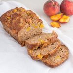 Sliced loaf of peach banana bread on a white cloth with some sliced peaches in the background