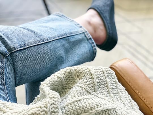 Close up of Shannon’s leg crossed over opposite knee sitting in leather chairing wearing denim and wool slippers with a textured grey knit in lap.