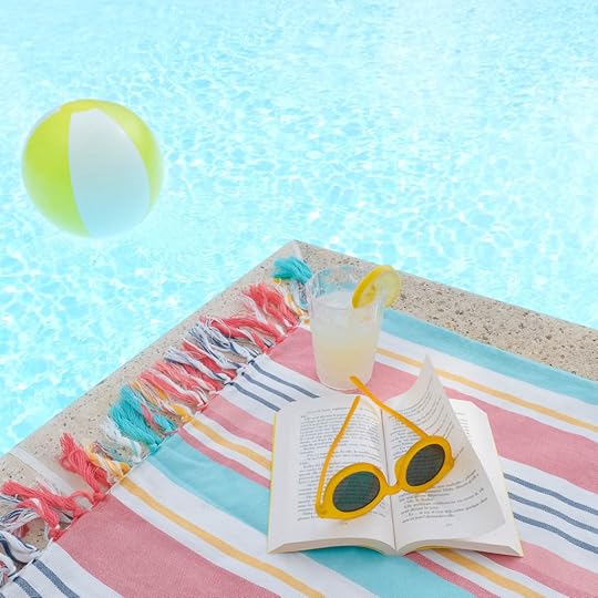 Striped beach towel on a pool deck with a paperback book open and yellow sunglasses on top of book. Yellow and white inflatable ball floating in pool.