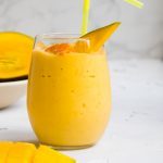 large glass filled with citrus mango smoothie with a yellow straw and some sliced mango in the background