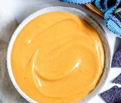 There's a gray bowl filled with creamy orange vegan nacho cheese. There are some chips on a white table beside the bowl, along with a colorful towel.