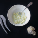 mixed garlic butter in a white bowl next to silver garlic press and head of garlic.