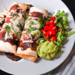 two bean and beef burritos on a white dinner plate topped with sauce, cheese, and cilantro.