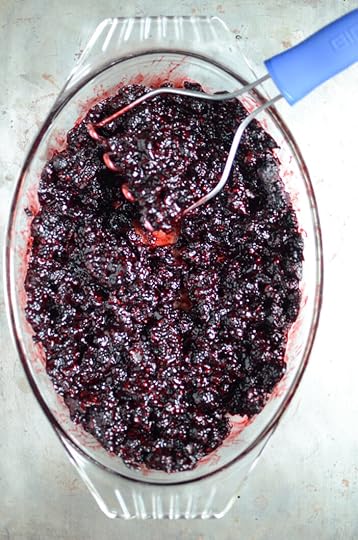 Smashed black raspberries in an oval glass baking dish, with a blue handled potato masher in the upper right hand corner.
