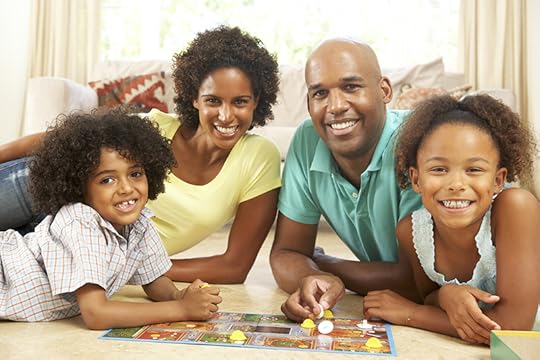 A family smiles at the camera as they play board games on the living room floor. Although times may be challenging, these back to school quotes from Thomas Mitchell remind us that there are always ways to find happiness.