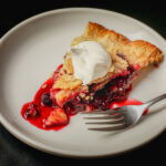 white dessert plate with a slice of mixed berry pie and a dollop of whipped cream and a fork.