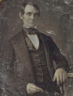 Lincoln, posed stiffly in coast, vest and bow tie, gazes at camera, left elbow propped on a book