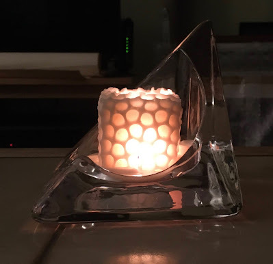 White soy candle, cubic, with a hive-patterned surface, the variegated surface illuminated by the flame inside.