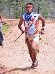 Tarahumara man with headband and pole, running on a brown dirt road with green trees in the background.