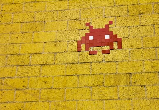 Image: street art of a pixelated video-game style space invader made of red tiles applied to a yellow brick wall.