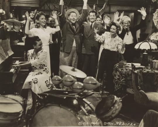 Scott, in a floral dress at piano, playing piano with a brilliant smile, turning to look at Red Skelton and other cast members, hands raised during a scene.