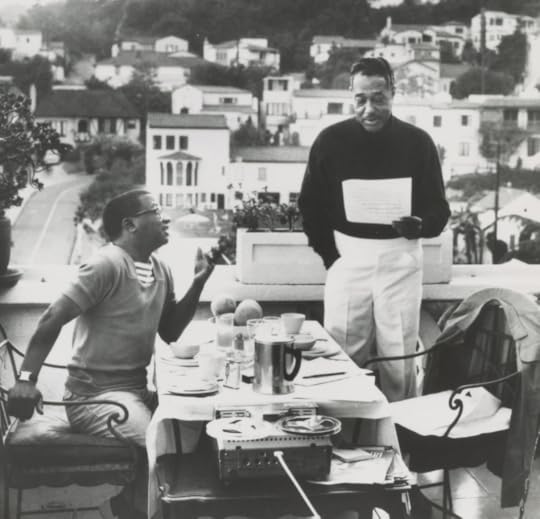 Strayhorn and Ellington, working with music at an open-air breakfast on a balcony, with houses on hills behind them. Breakfast plates and coffee pitcher on table. Reel to reel tape recorder beside table.Strayhorn sitting, gesturing with left hand. Ellington, standing, holding sheet music in left hand, right hand in pocket of his slacks.Photo is black and white. 