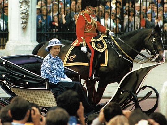 Photo of Queen Elizabeth riding in an open carriage. Beside her a uniformed member of the military rides on horseback.